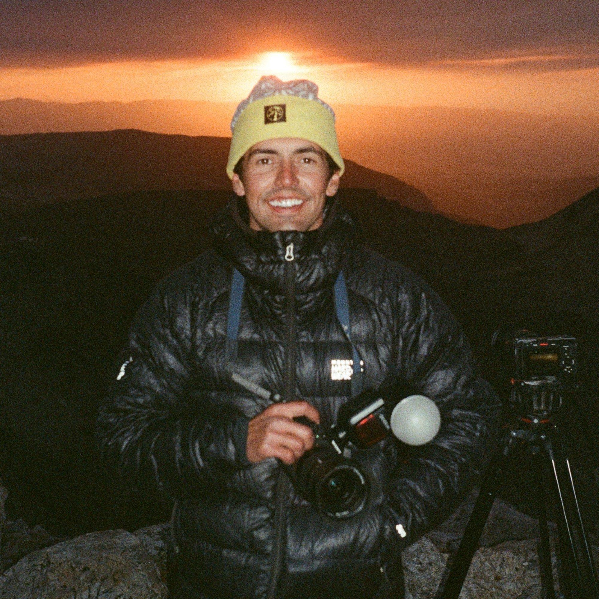 A tripod pointed at the sunset behind him, professional mountain athlete and photographer Clayton Herrmann gives a candid smile.