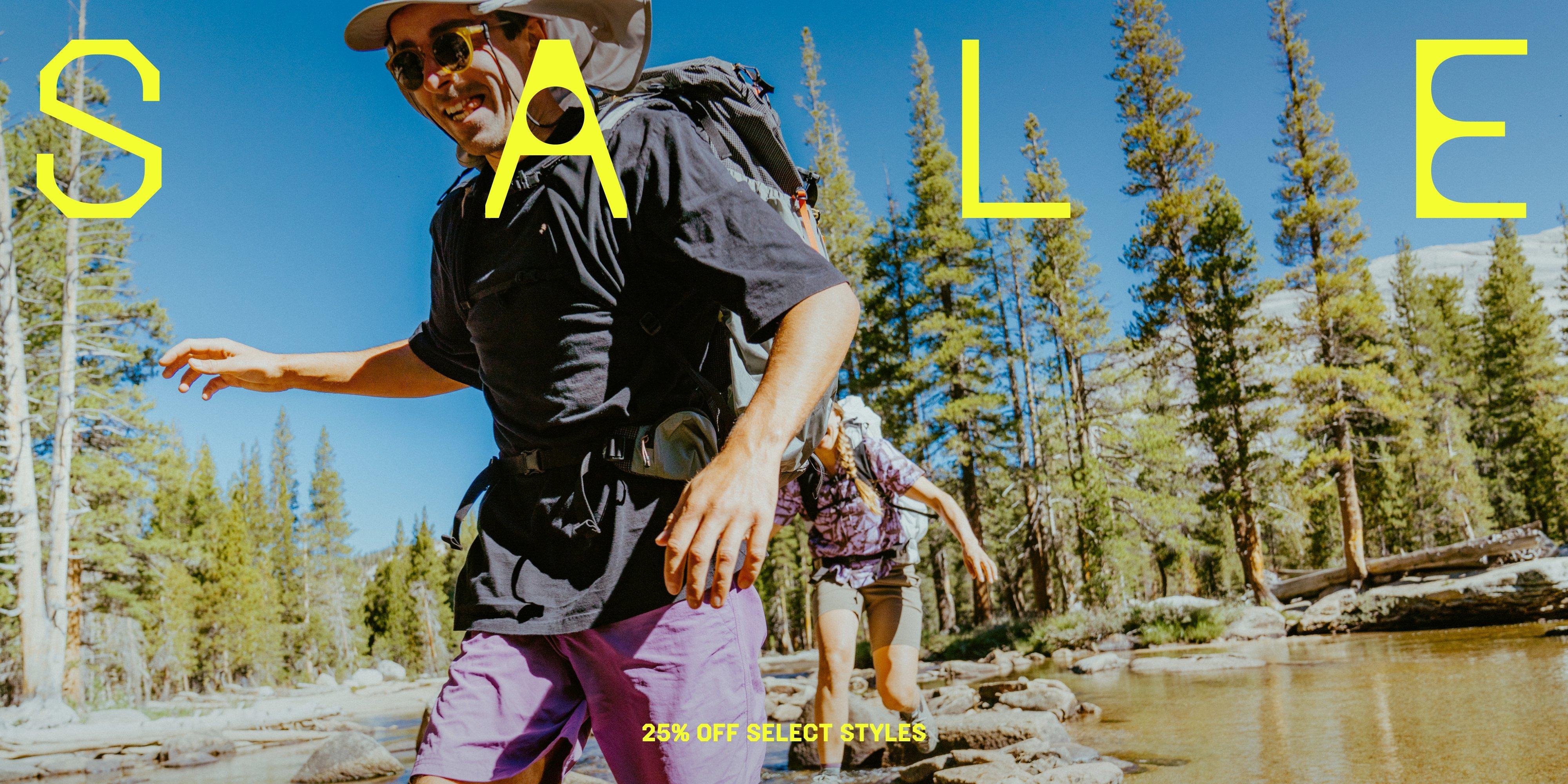 A man in Stryder™ Shorts and woman in a Sun Drift™ Cooling Shirt hop along rocks to cross a river while backpacking.