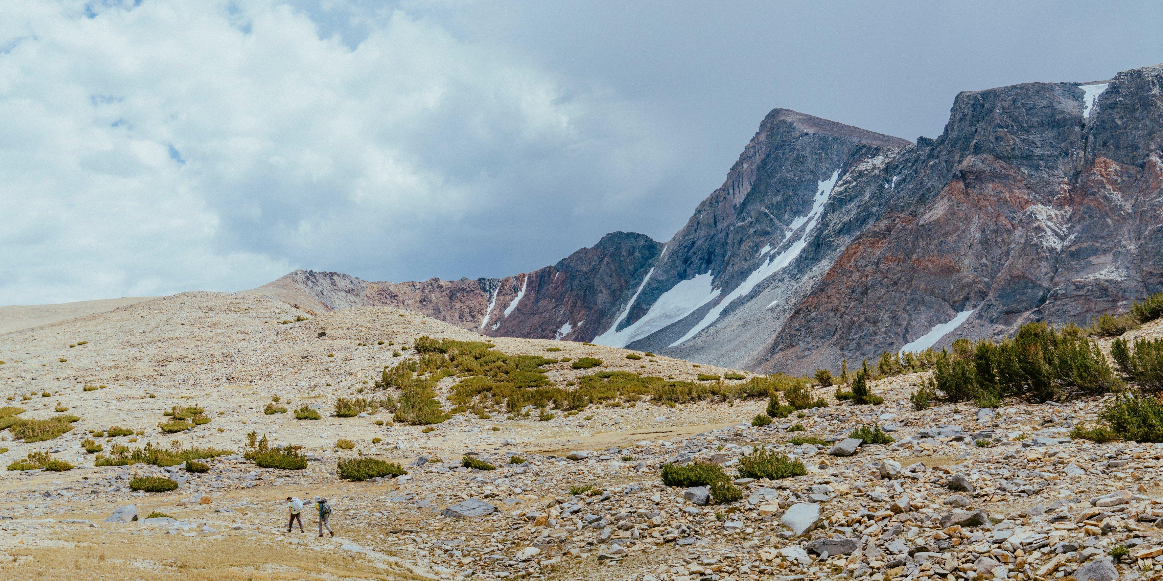 Two seemingly tiny backpackers traverse a rocky field with foggy peaks dappled in snow loom in the background.