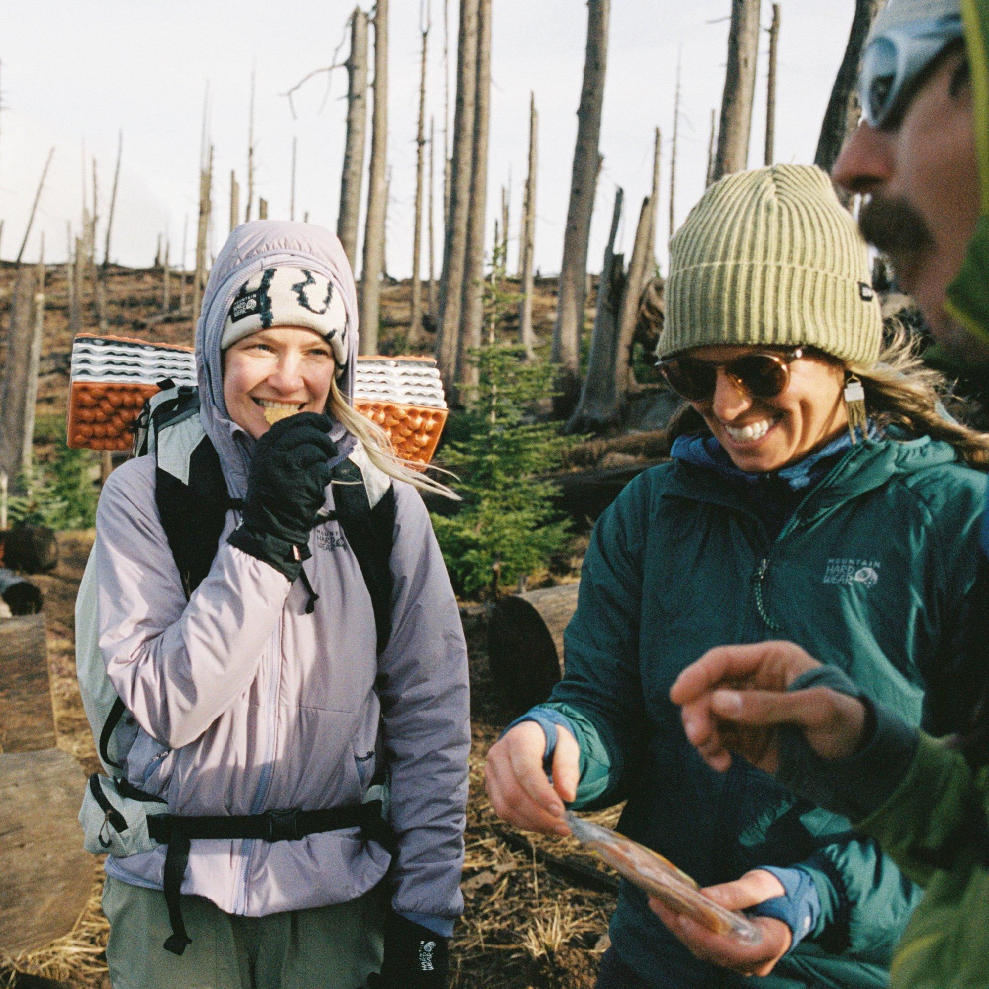 Three backpackers in Kor Alloy™ Jackets stop to snack on dried fruit, barren trees behind them and wind in their hair.