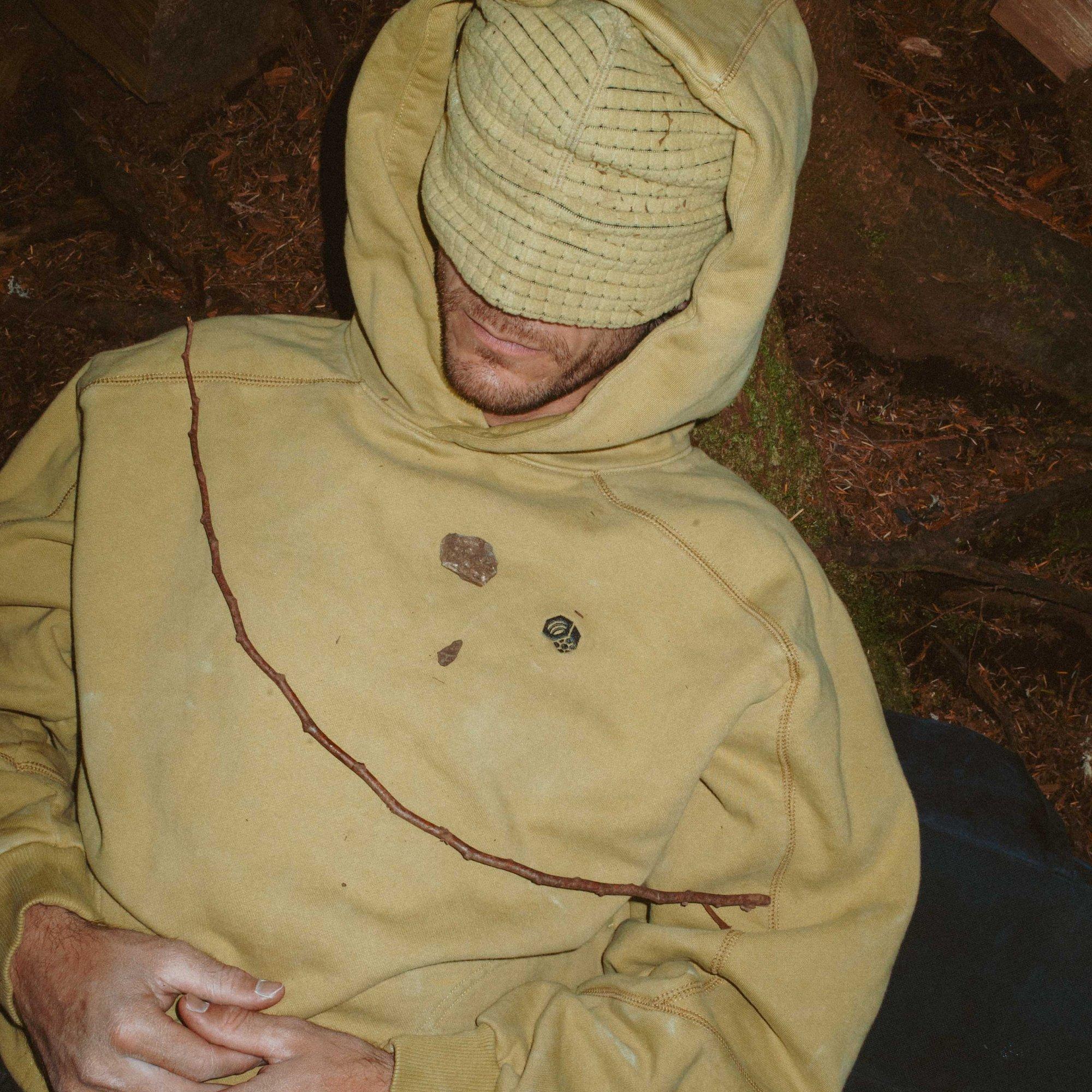 A man in a chalk-dusted Hardwear Supply™ Hoody snoozes against a tree. Rocks and branches on his chest form a happy face using the nut logo as one eye.