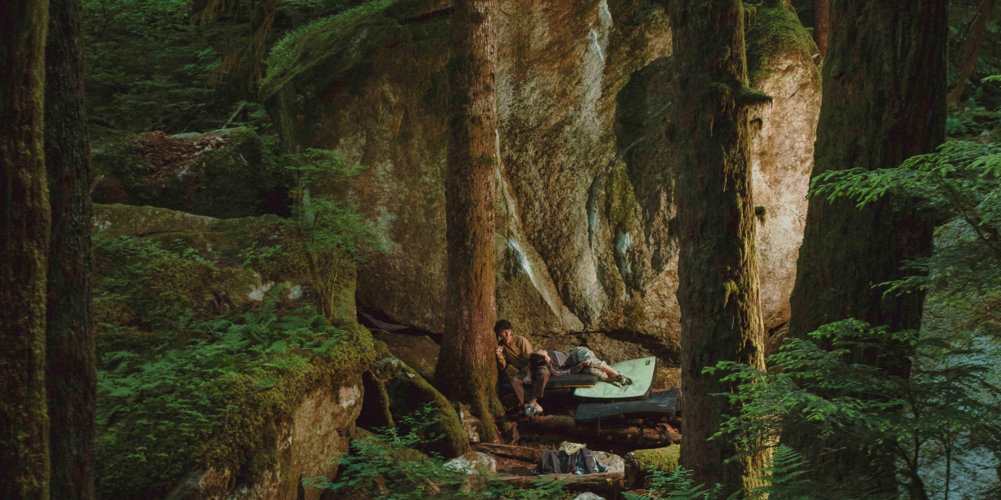 A male and female climber lounge on crash pads at the base of an immense rock face framed by mossy trees.