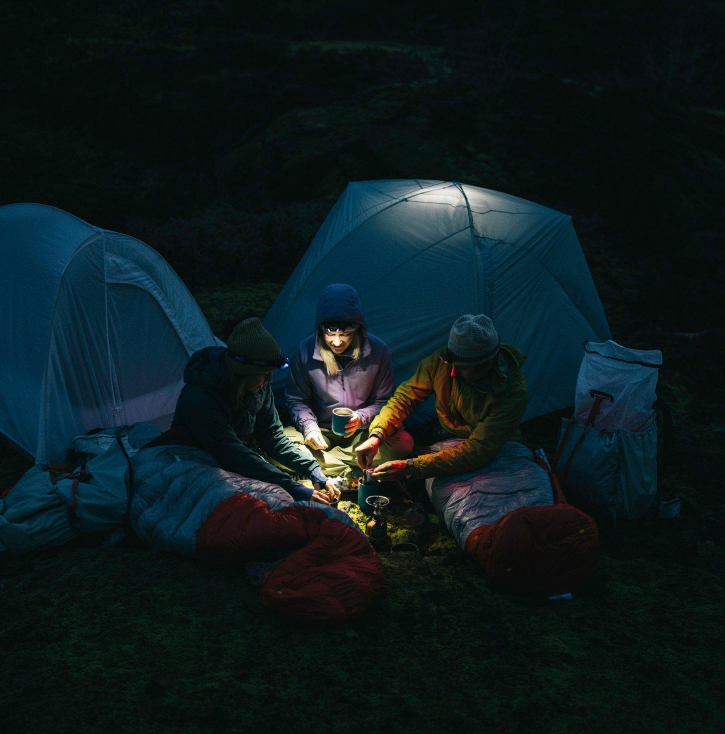 Illuminated by headlamps, three people make coffee in front of their tents.