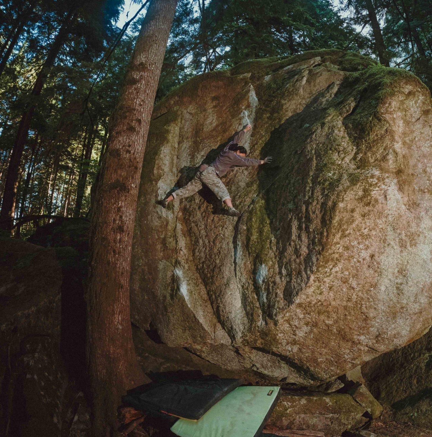 A boulderer wearing Hardwear Supply™ finds a toehold on a large, mossy boulder face. 