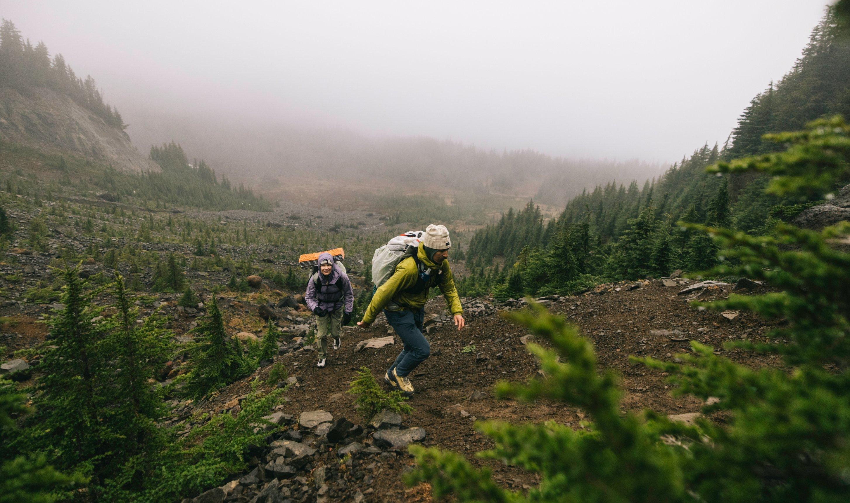 Two backpackers in StretchDown™ Hooded Jackets ascend groomed trail into the treeline. 