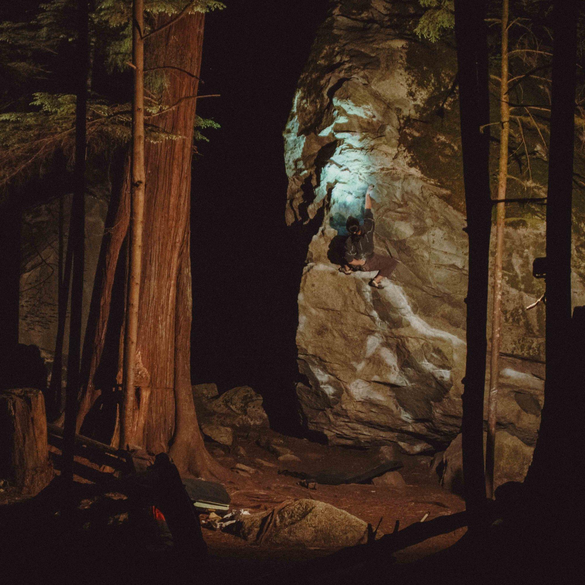 Framed by shadowy trees, MHW Athlete Rell Lennox reaches for her chalk while bouldering, her headlamp illuminating her next move.
