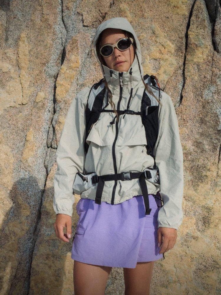 A woman with hiking pack wearing a Stryder Jacket and Dynama Skort in front of a rock face.


