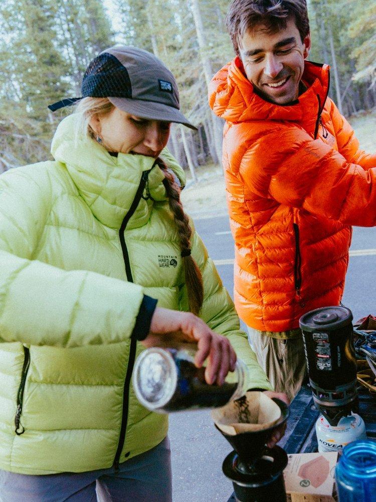 A woman and man wearing brightly colored, puffy down jackets make coffee at camp.